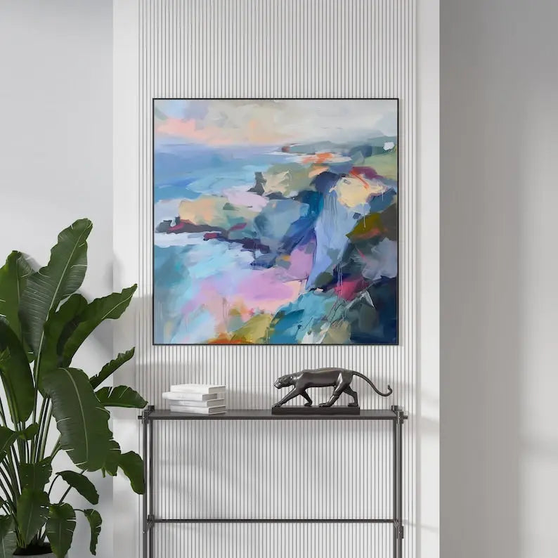 A large square painting featuring the blue sea framed by blue and cyan mountains, enhanced by hues of pink and orange. 
