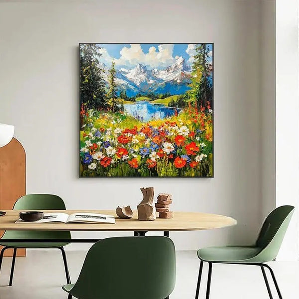 A large square painting of a beautiful lakeside landscape bordered by colorful flowers in red, white and blue here while snowy mountains rise on the horizon. 