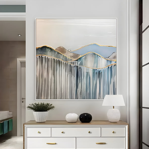 An abstract square painting of a mountain scene with layered blue and beige forms, golden accents, and a horizon that suggests stillness and depth. 