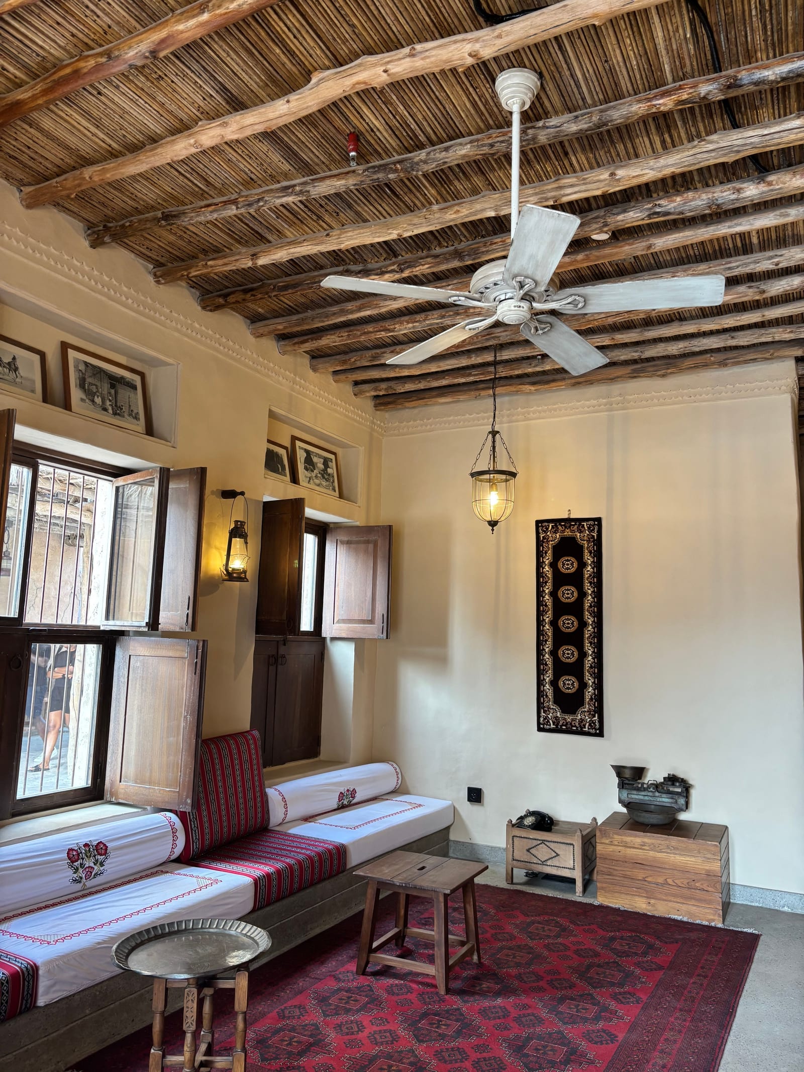 A traditional Gulf sitting room with a palm-beam ceiling, low cushioned bench seating, red carpet and framed prints on white walls.