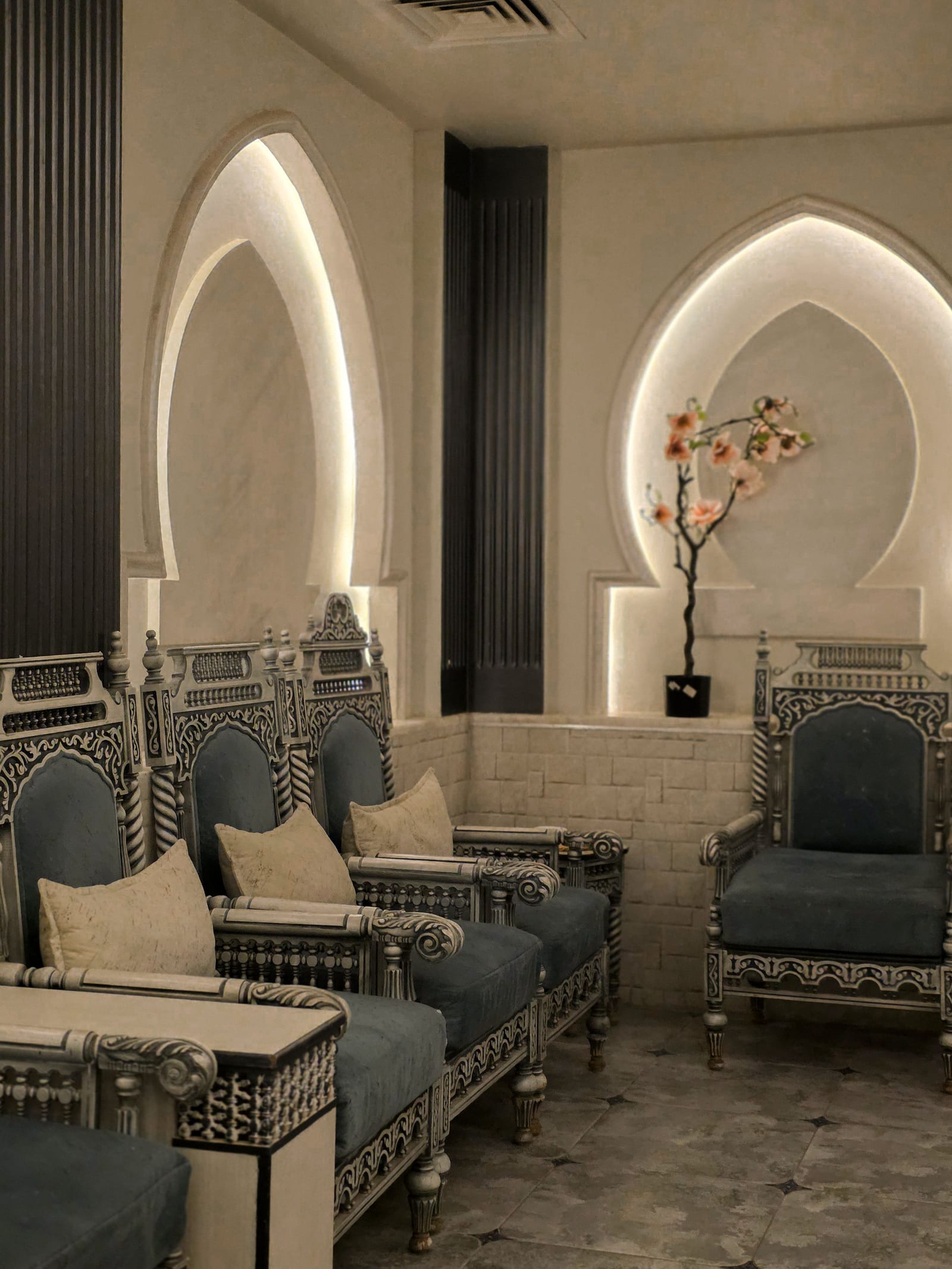 A sitting room with backlit pointed-arch niches, cream stone walls and a row of carved chairs in teal upholstery.