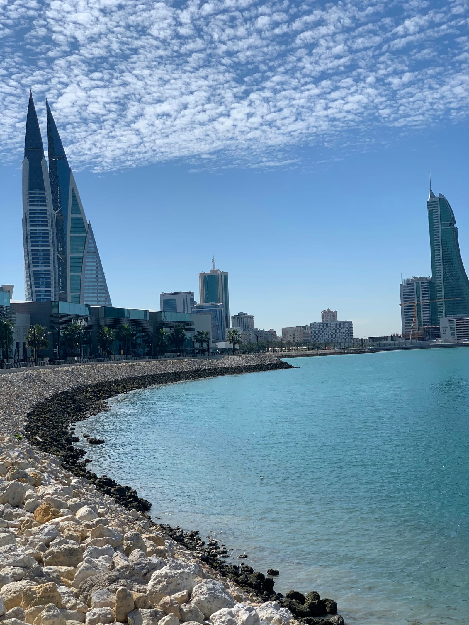 The sail-shaped Bahrain World Trade Center and the Harbour towers over turquoise water and a rock-lined shore, Bahrain Bay, January 2021.