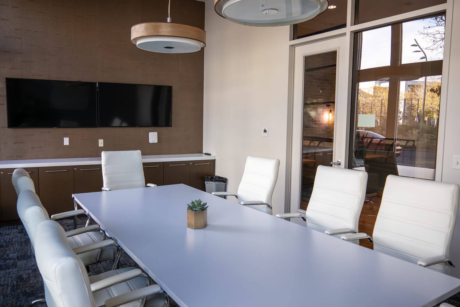 A modern boardroom with a long white table, white leather chairs and two screens on a dark textured feature wall.