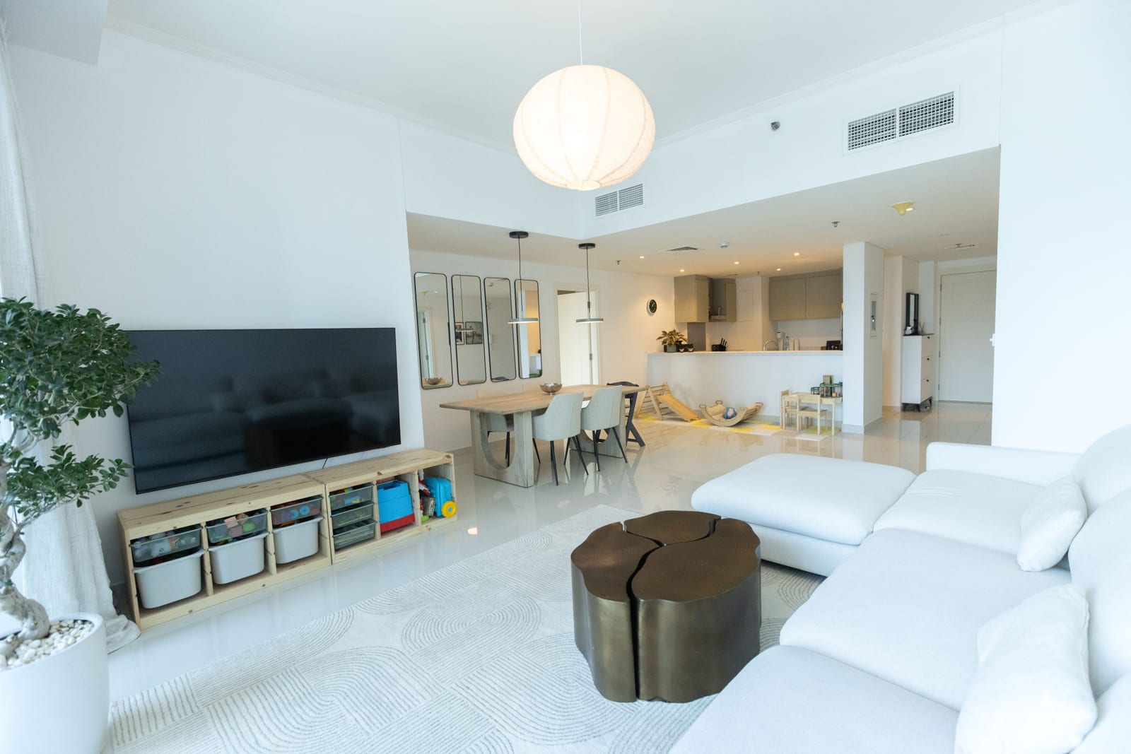 A white open-plan Gulf apartment living room with a marble floor, cream sectional sofa, television wall and dining area behind.
