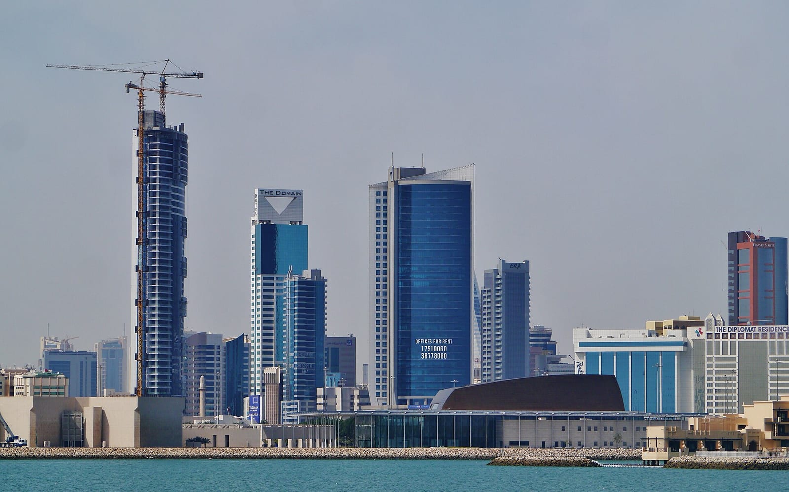 The Bahrain National Theatre with its gold latticed canopy at the waterline, with the Manama towers and a crane behind it.