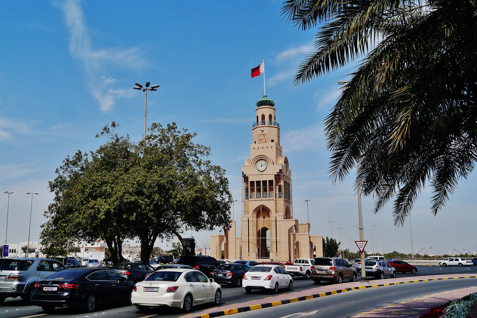 The Riffa clock tower with a Bahraini flag, palms and traffic on the roundabout in front of it.
