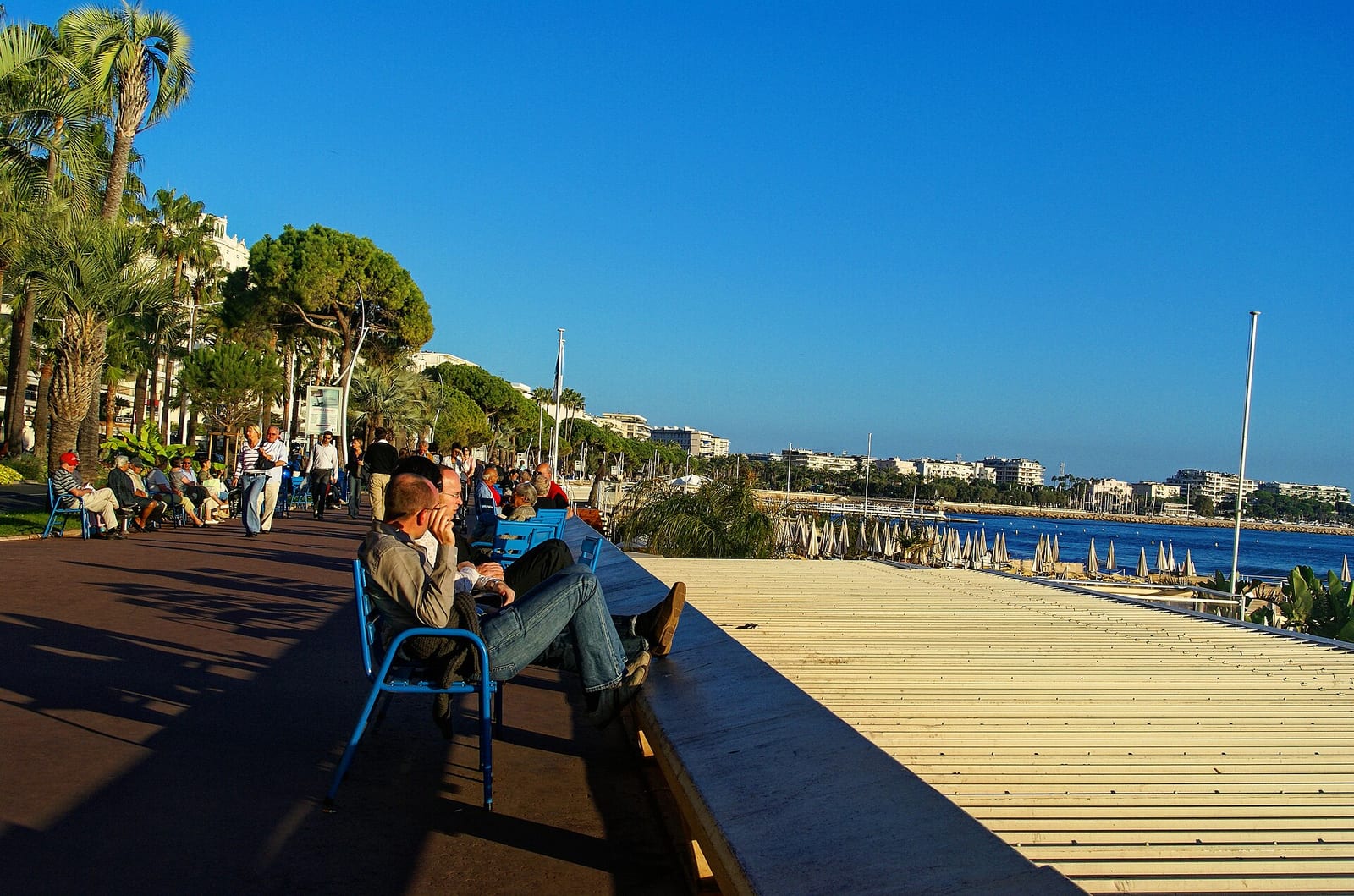People sitting on benches along the Boulevard de la Croisette in Cannes under palm trees with the bay beyond.