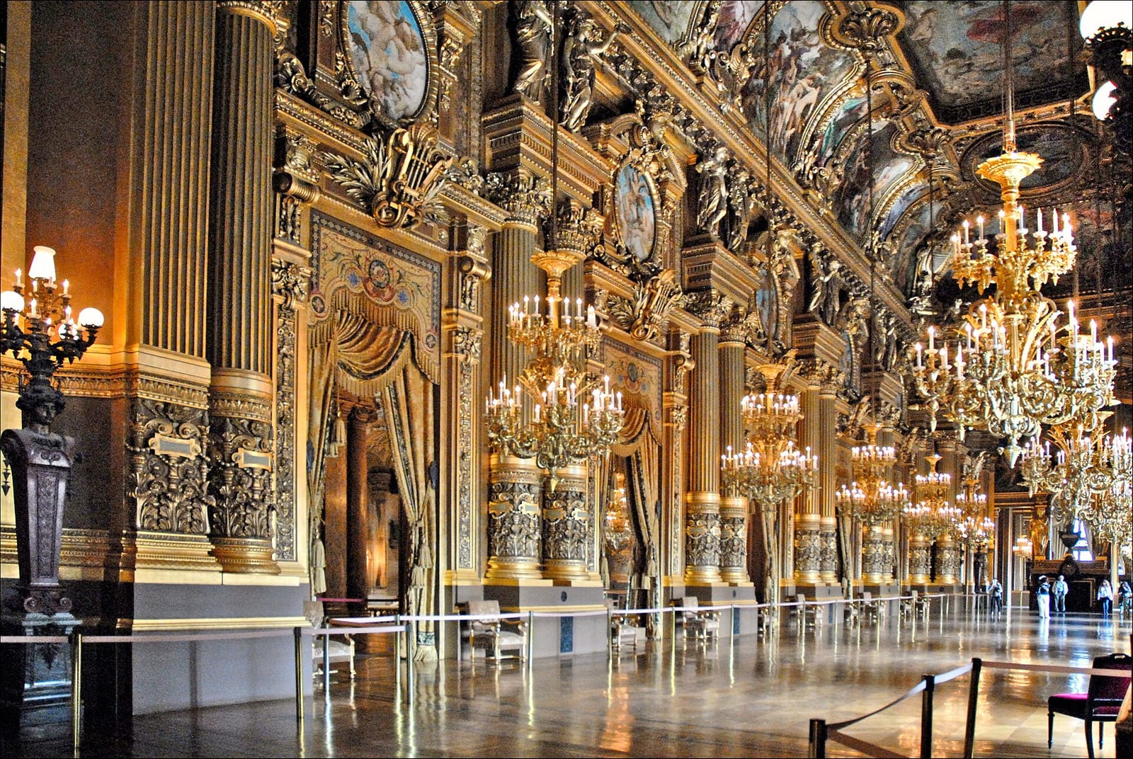 The gilded grand foyer of the Palais Garnier in Paris with chandeliers, painted ceilings and mirrors.