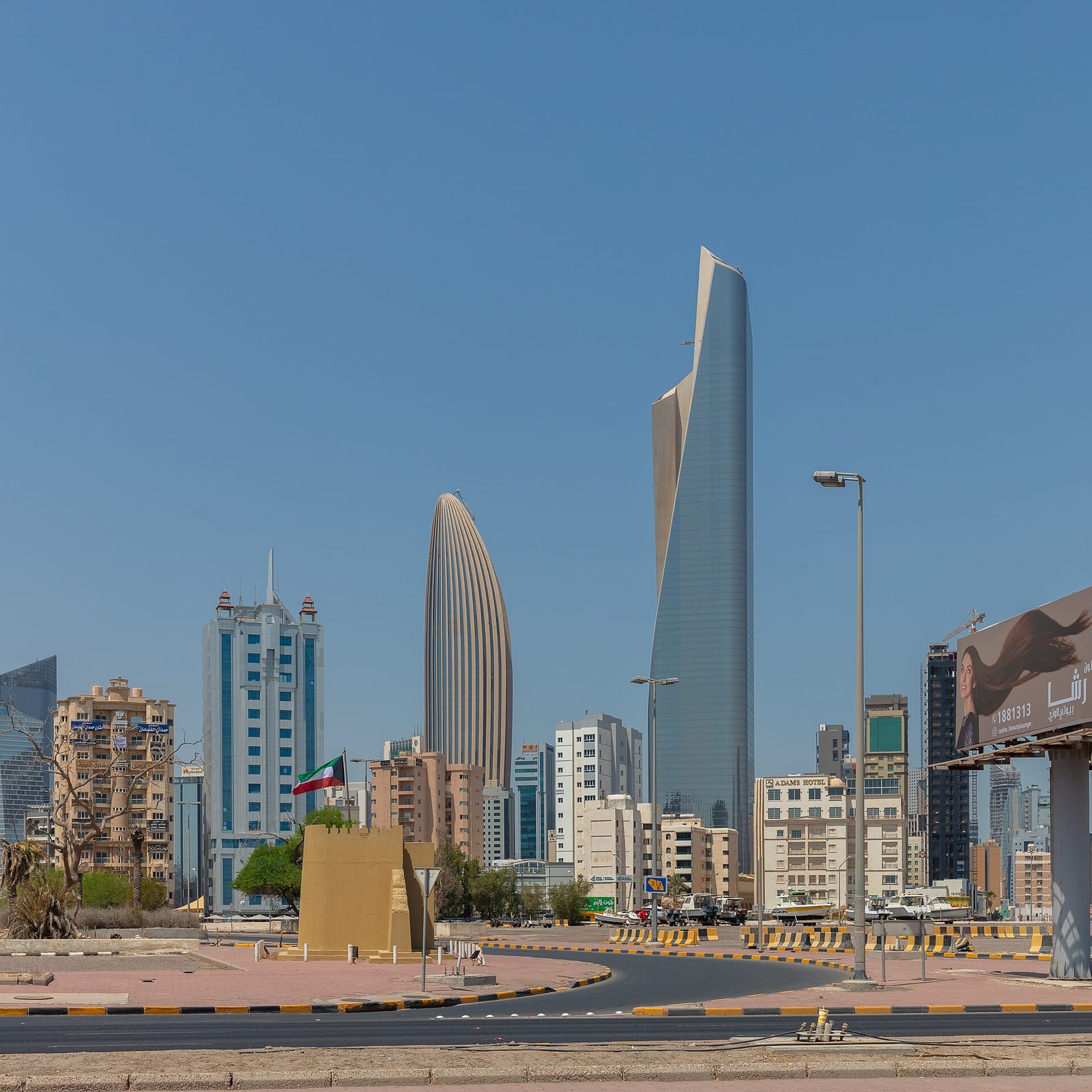 Square-format view of glass and stone office towers in downtown Kuwait City against a hazy August sky, 2024.