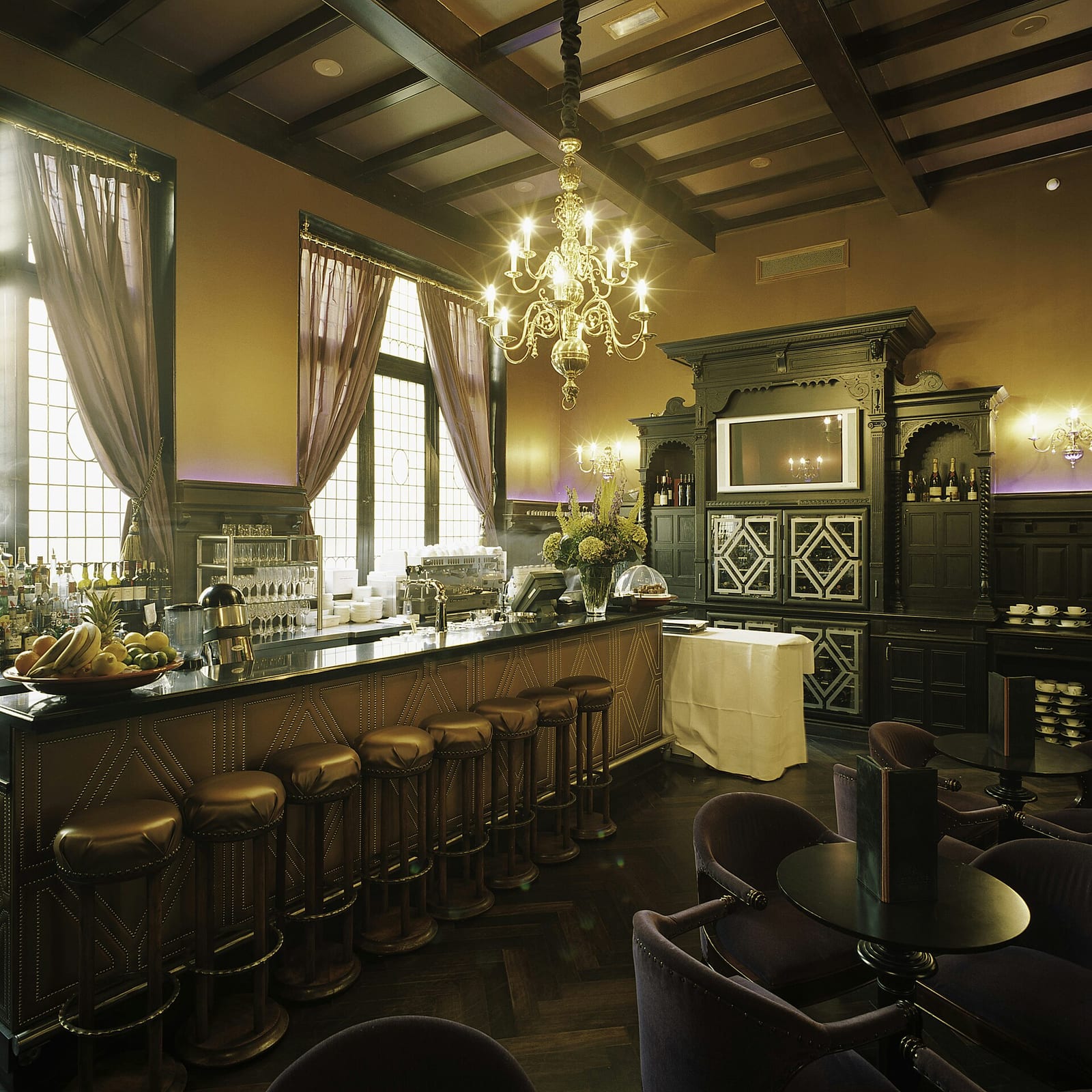 The wood-panelled bar of Hotel Des Indes in The Hague with leather stools, a chandelier and a mirrored back bar.