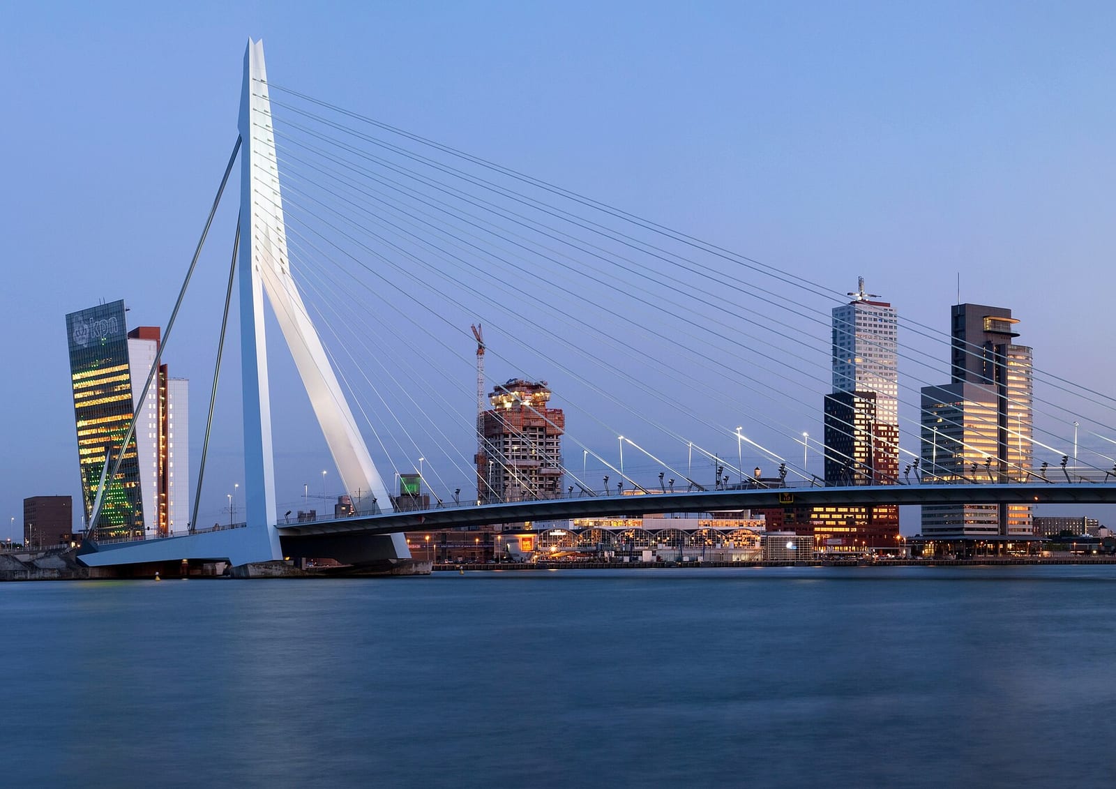 The white Erasmus Bridge over the Nieuwe Maas in Rotterdam at dusk, with the Kop van Zuid towers behind it.