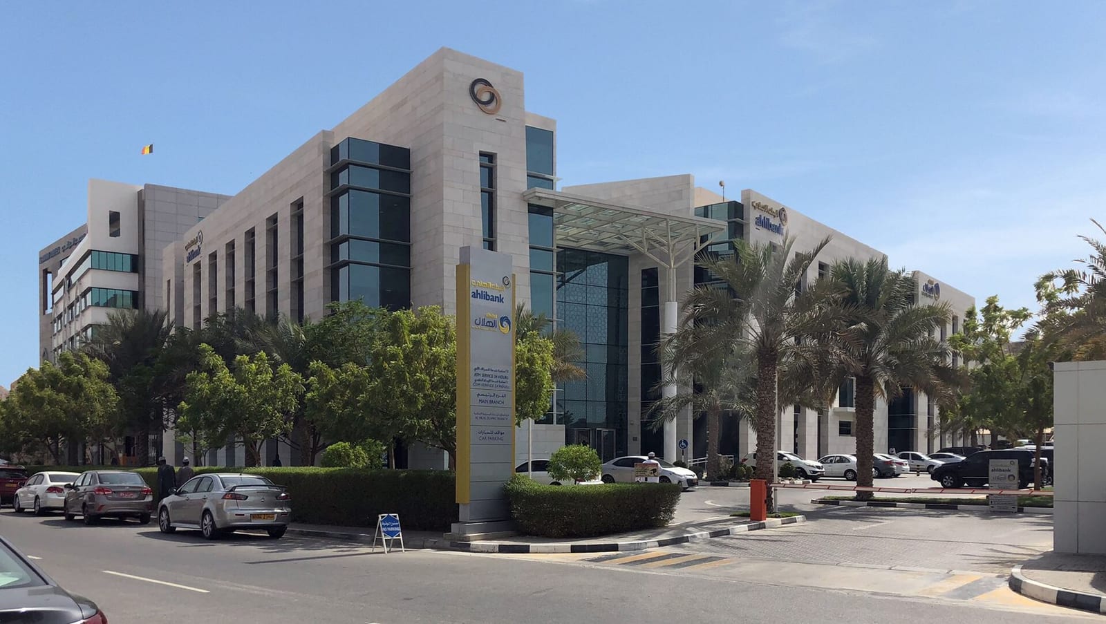 Ahlibank head office in Muscat, a cream-and-grey contemporary building with a round tower, palms and a car park in front.