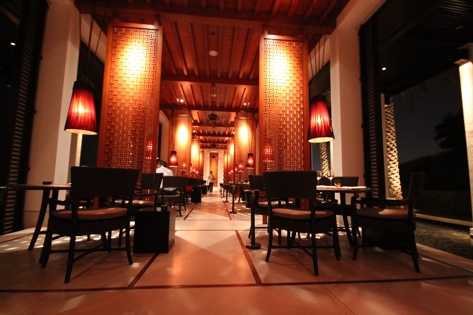 Dimly lit dining room of The Beach restaurant at The Chedi Muscat, with dark timber, lantern light and a long central aisle.