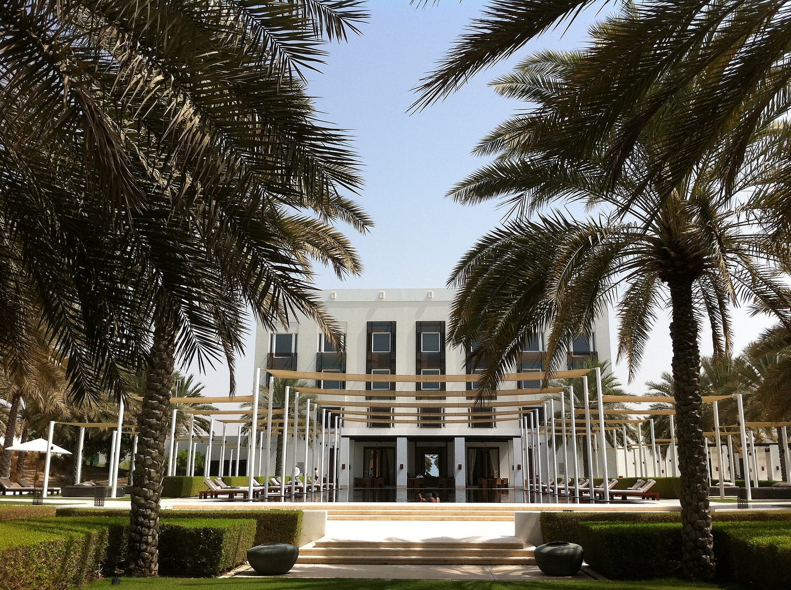 Low white pavilions of The Chedi Muscat behind palms and clipped lawns, with the Hajar mountains in the background.