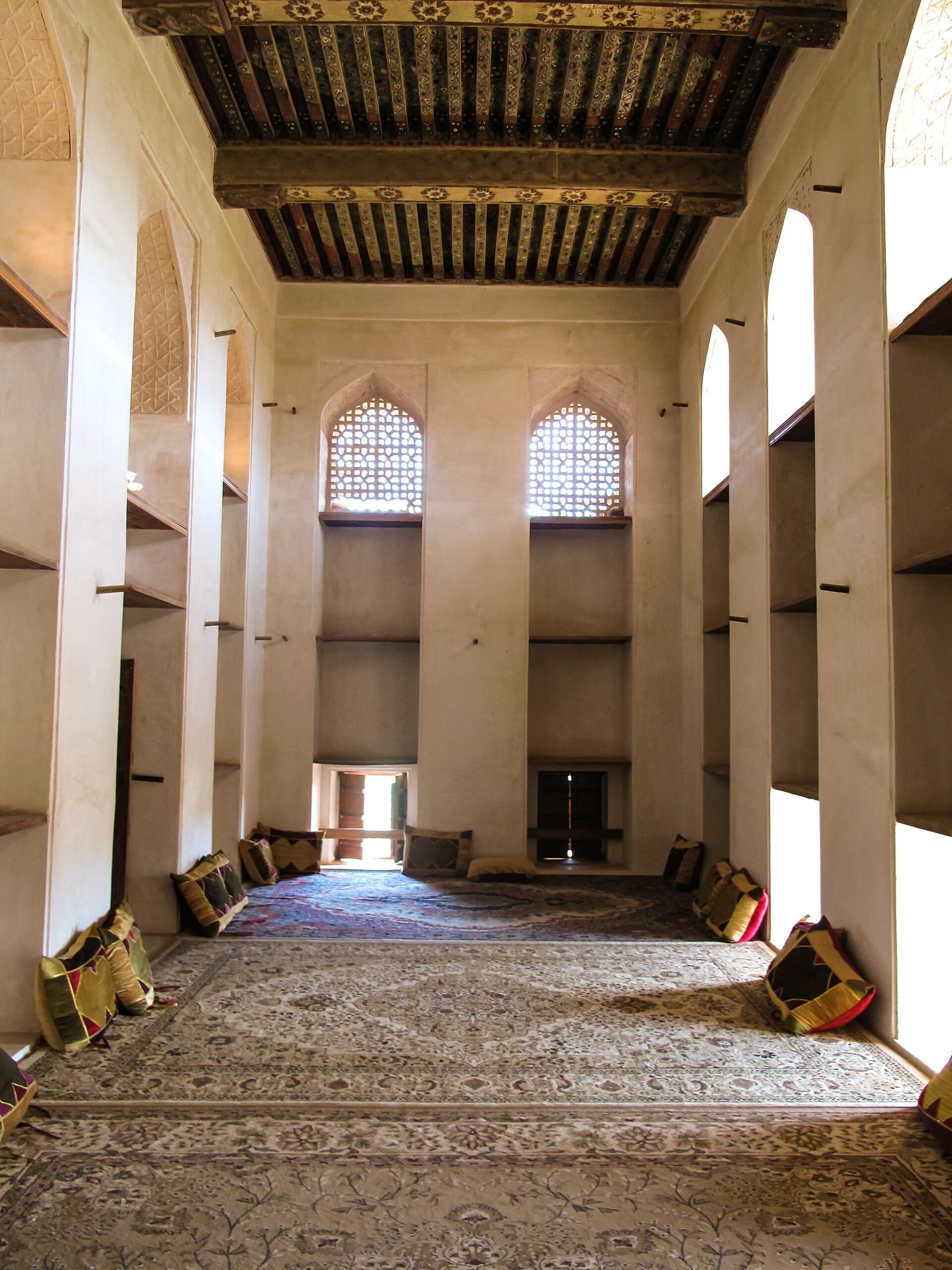 Long reception hall inside Jabrin Castle, Oman, with floor cushions along both walls, arched niches and a painted timber ceiling.
