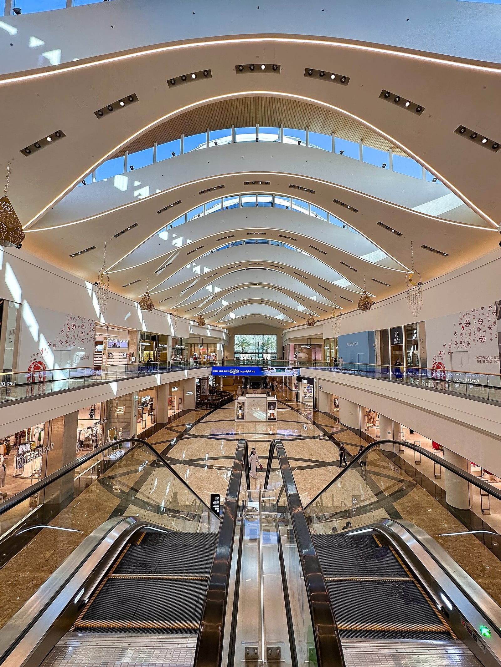 Vaulted white atrium of the Mall of Oman in Muscat with escalators, glass balustrades and a long suspended ceiling.
