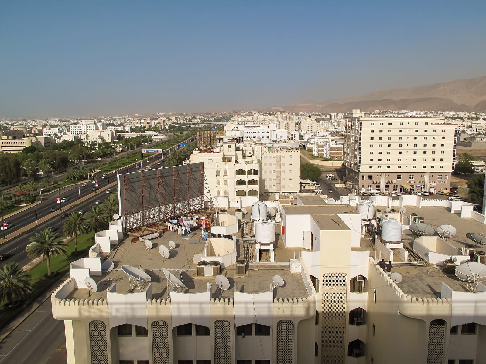Rooftops of white villas and mid-rise blocks in Muscat's western suburbs, seen from above with the coast beyond.