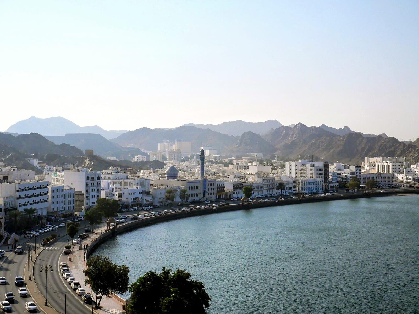 Aerial view of the Muttrah Corniche and harbour in Muscat, white low-rise buildings curving along the bay beneath bare rock hills.