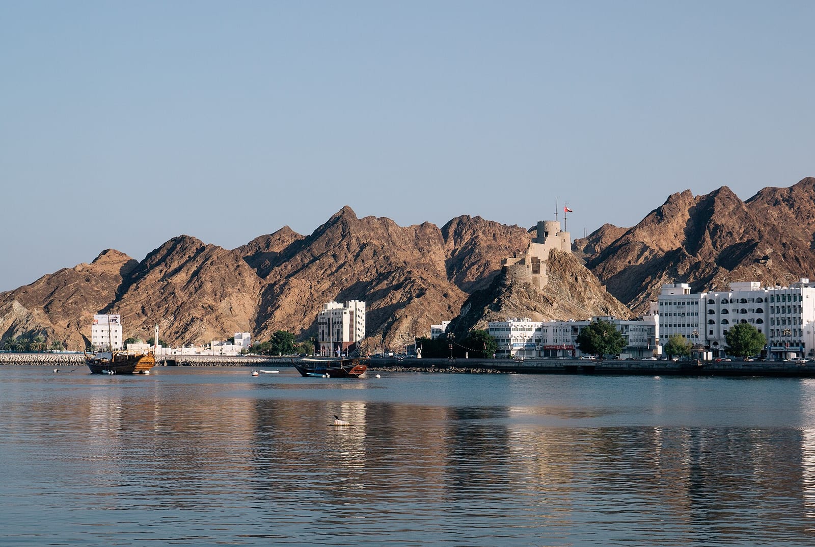 Muttrah Fort on a rocky ridge above the harbour and the white waterfront buildings of Muttrah, Muscat, seen from the Corniche.