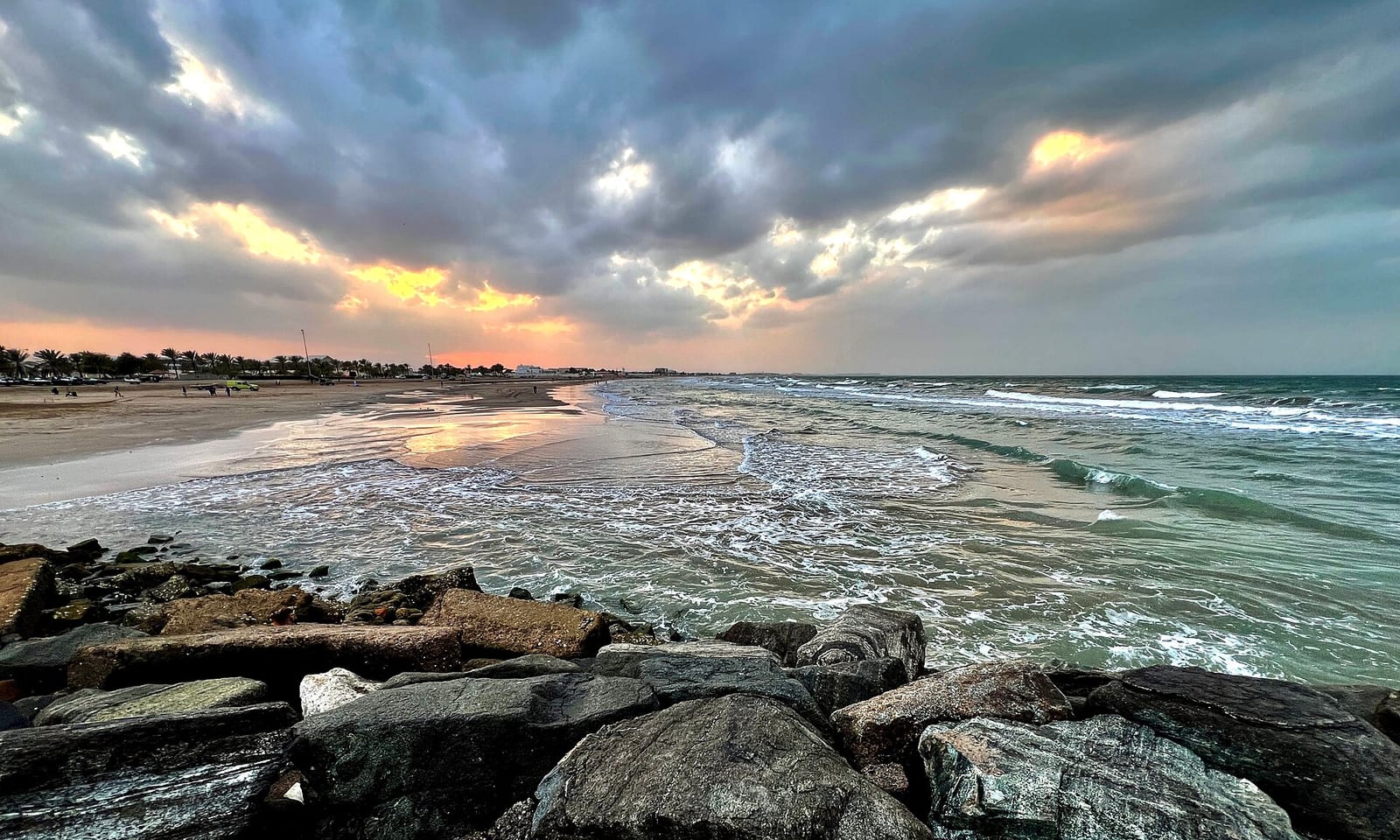 Dramatic evening sky over the surf at Qurum Beach, Muscat, with wet rocks in the foreground and long clouds catching the light.
