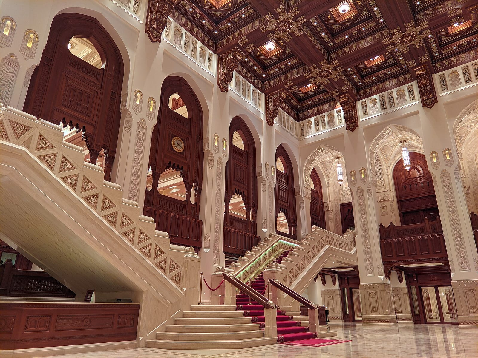 Marble foyer of the Royal Opera House Muscat with a grand staircase, pointed arches, carved timber ceilings and warm stone.