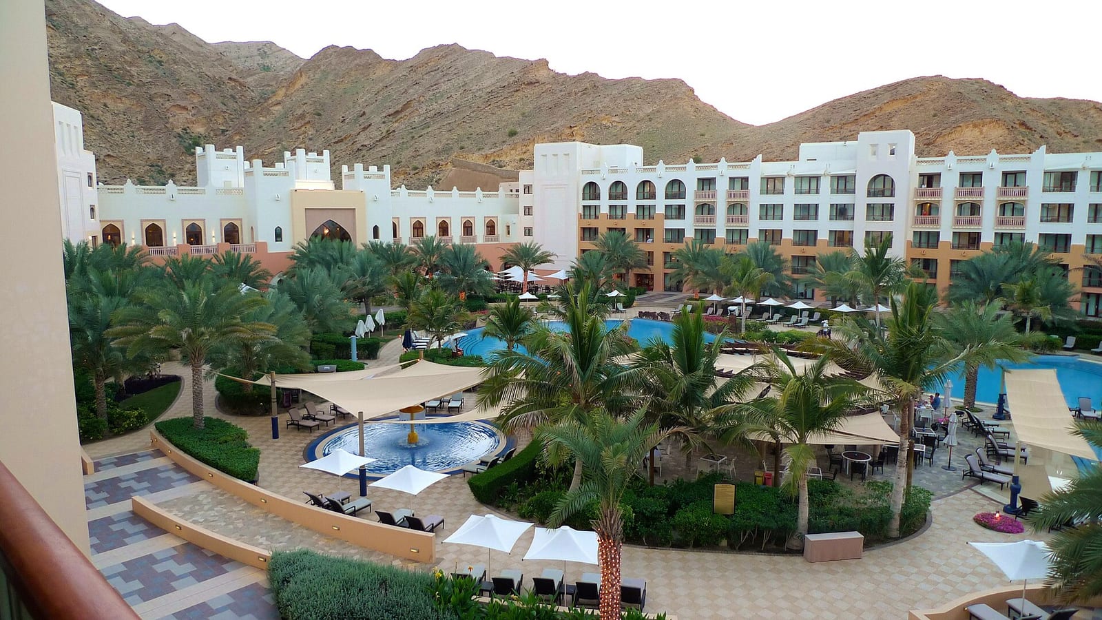 Shangri-La Barr Al Jissah resort in Muscat seen from a balcony: arched Omani-style wings around pools, lawns and palms under rock cliffs.