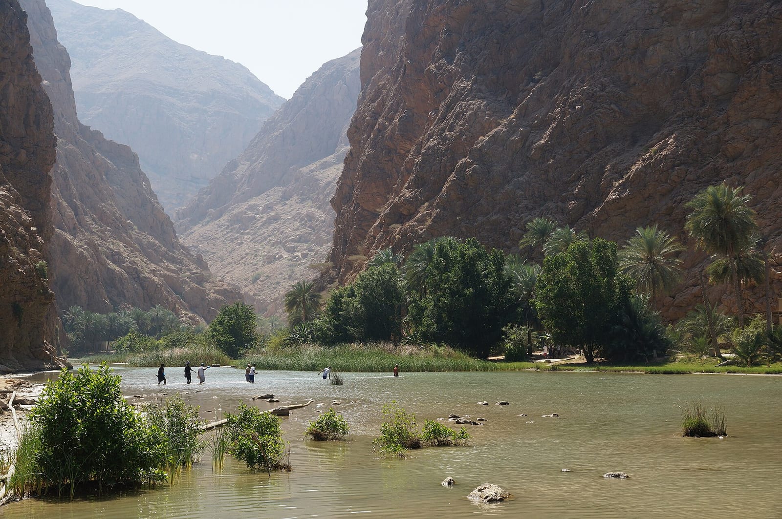 Wadi Shab in Oman: turquoise pools and palms between sheer grey rock walls, with small figures on the water's edge.
