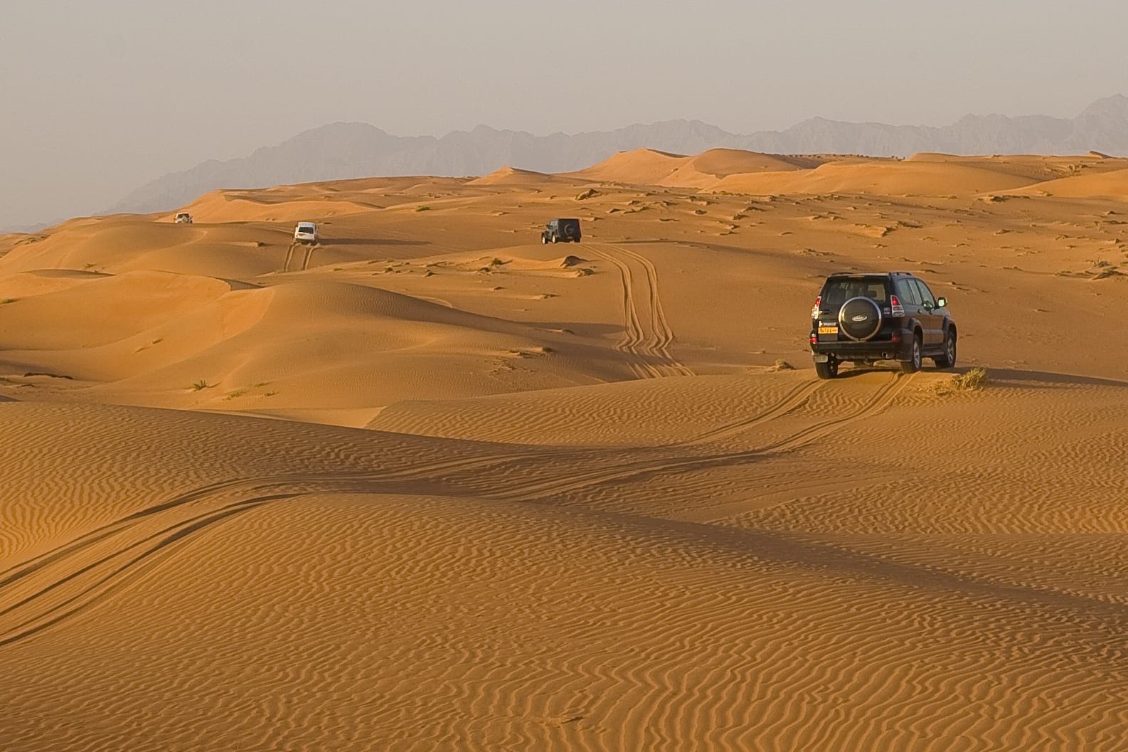 Wide view of orange dunes in the Wahiba Sands, Oman, with a single four-wheel-drive crossing the sand under a pale sky.
