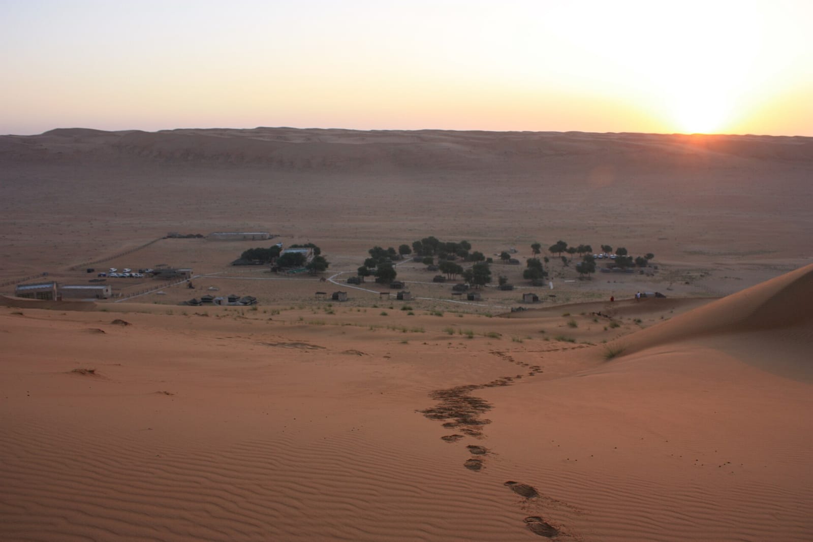 Sunrise over the Wahiba Sands in Oman, low light on rippled dunes and a line of footprints leading towards distant palms.