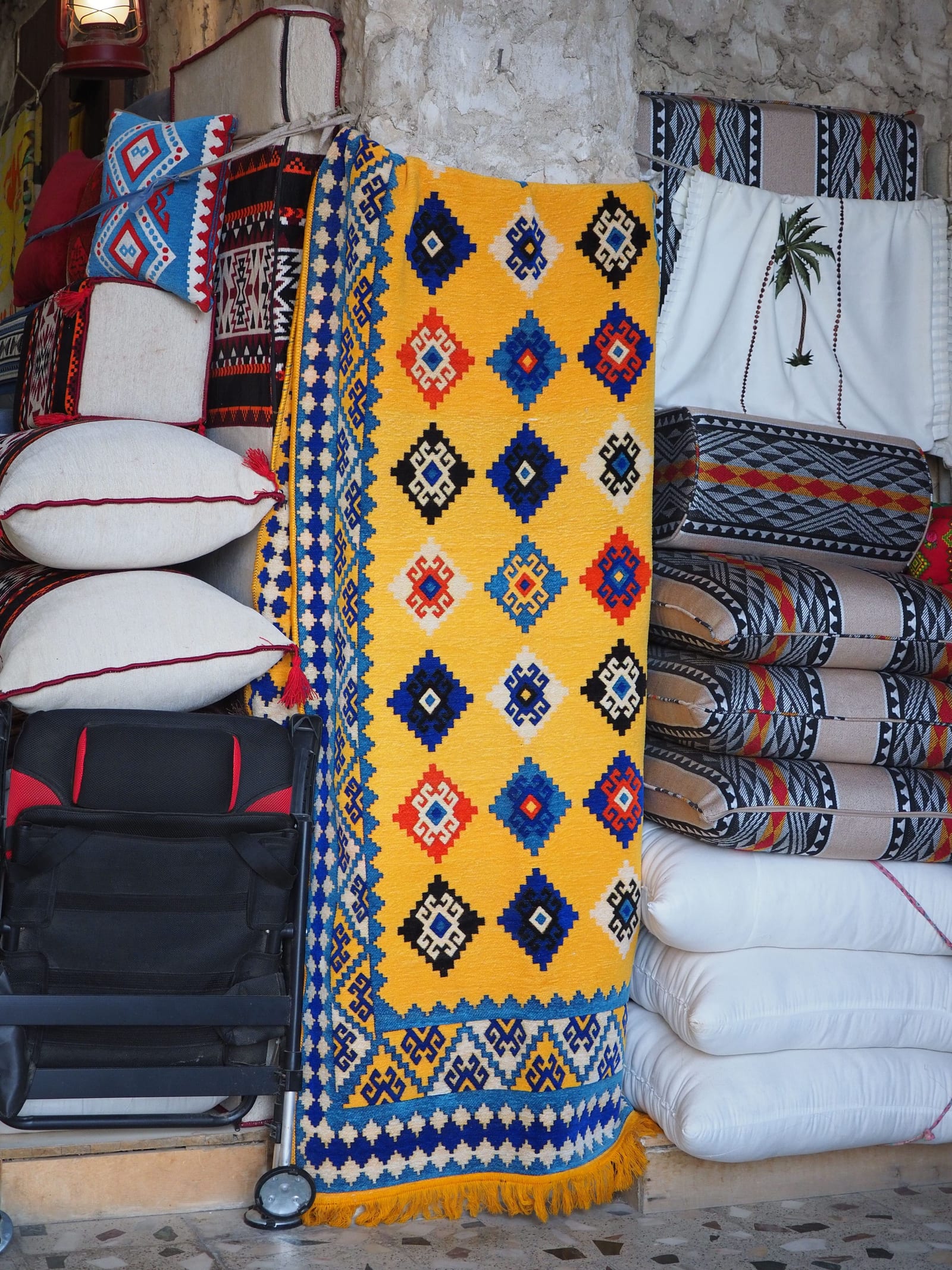A Doha souq stall stacked with sadu-pattern cushions and a yellow, blue and red diamond-motif woven rug.