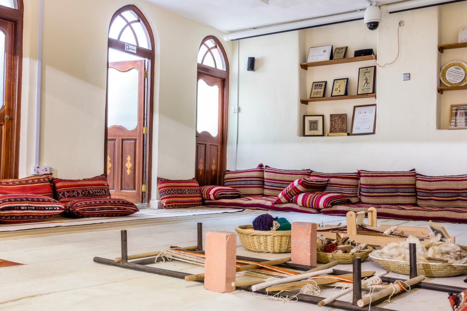 Sitting room with floor cushions in red sadu-style textiles along the walls, small wall shelves and weaving tools on the floor.
