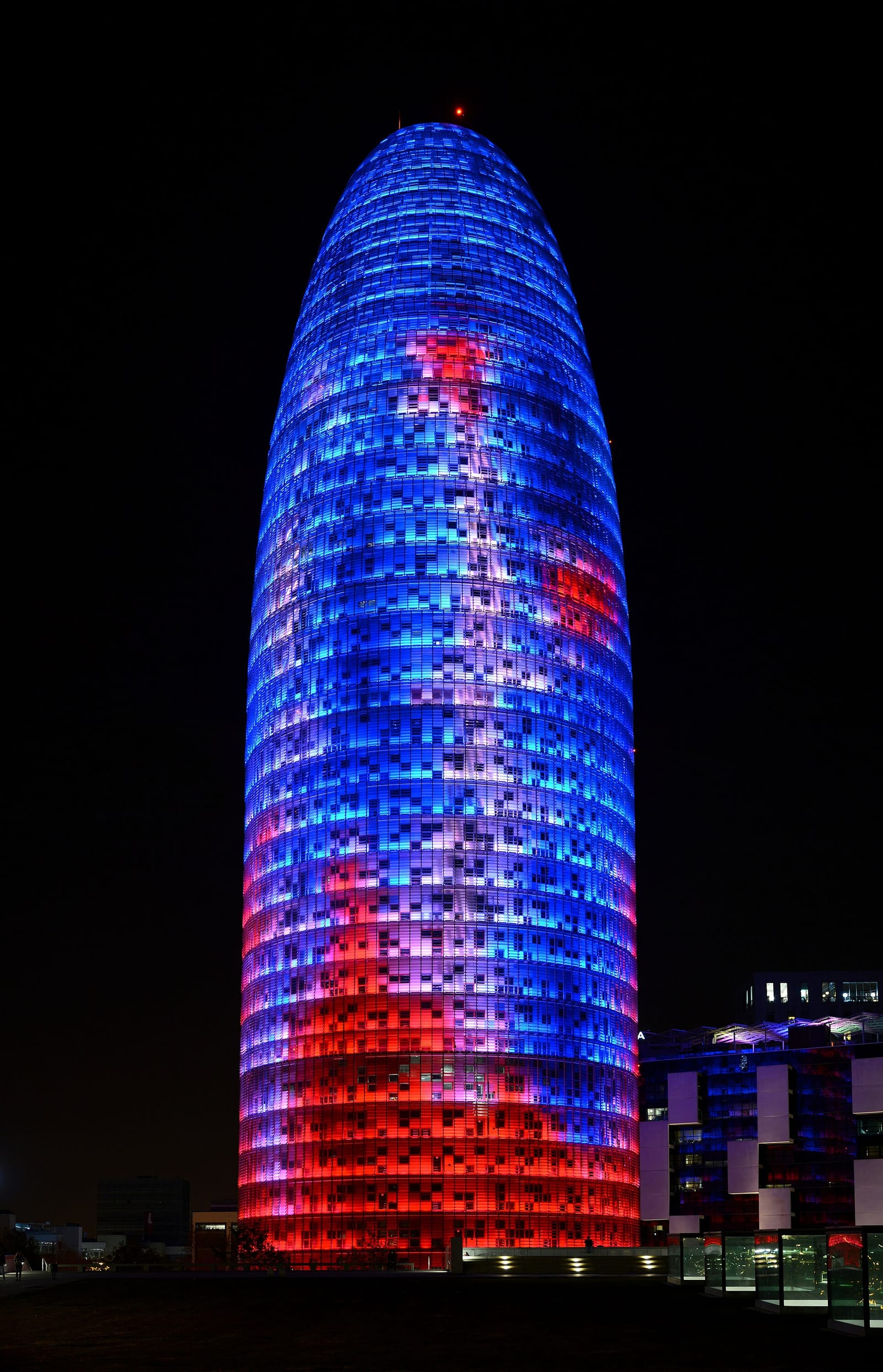 Torre Glòries in Barcelona at night, the bullet-shaped tower lit in blue and red against a dark sky.