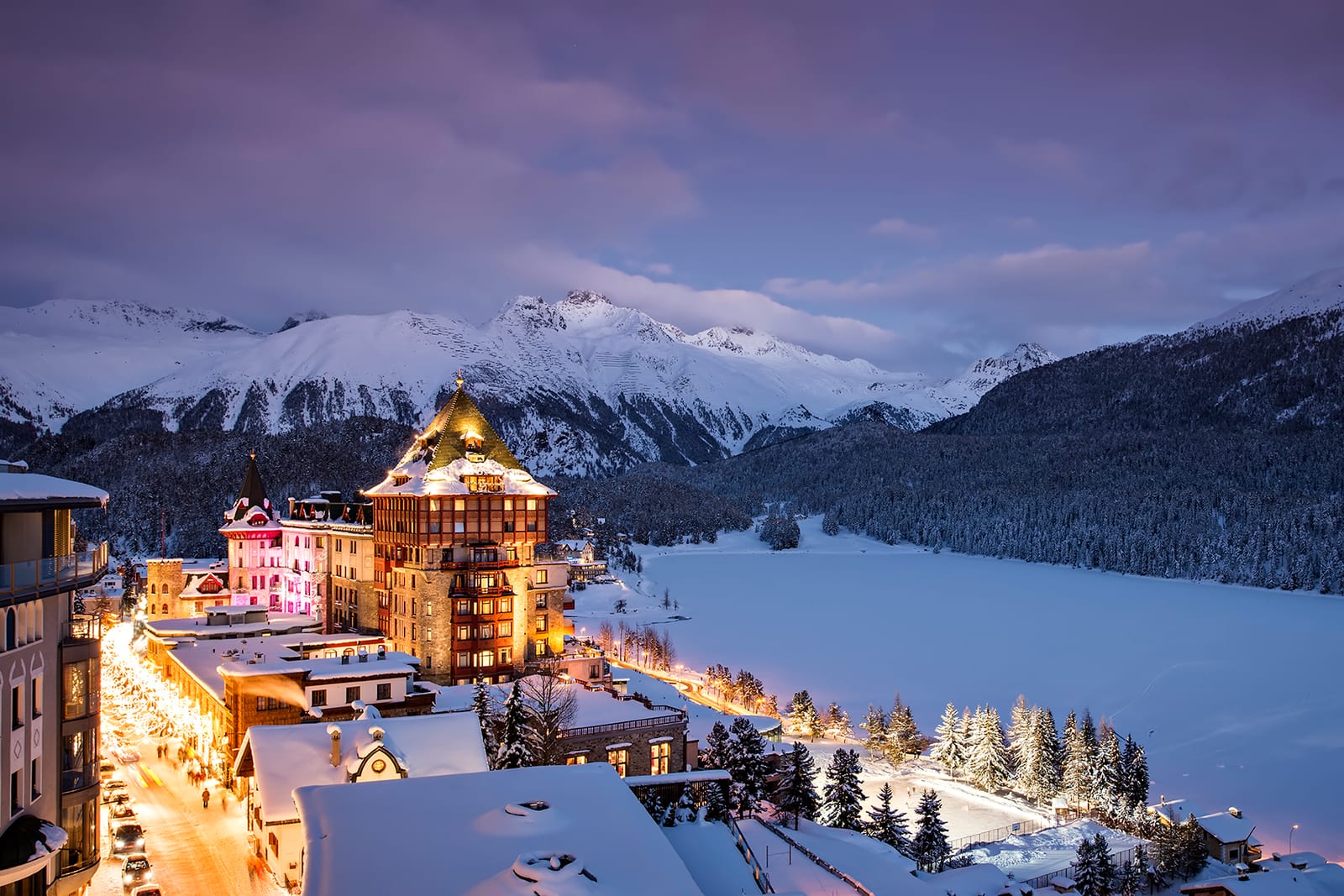 Badrutt's Palace Hotel in St. Moritz lit at dusk under snow, with the frozen lake and mountains beyond it.