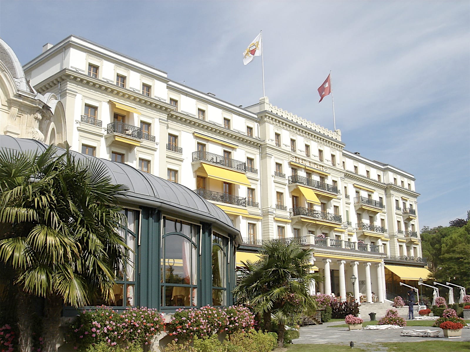 The white Belle Époque façade of the Beau-Rivage Palace in Lausanne-Ouchy with palm trees and flags in front.