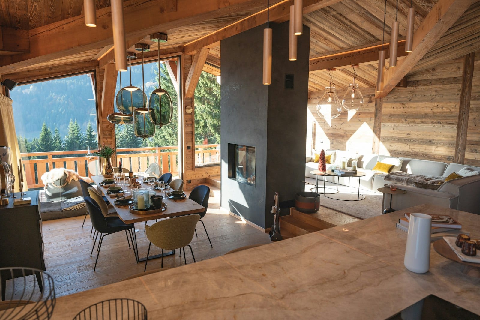 An open-plan chalet interior with timber beams, a dark fireplace wall, dining table under pendant lights and mountain views.