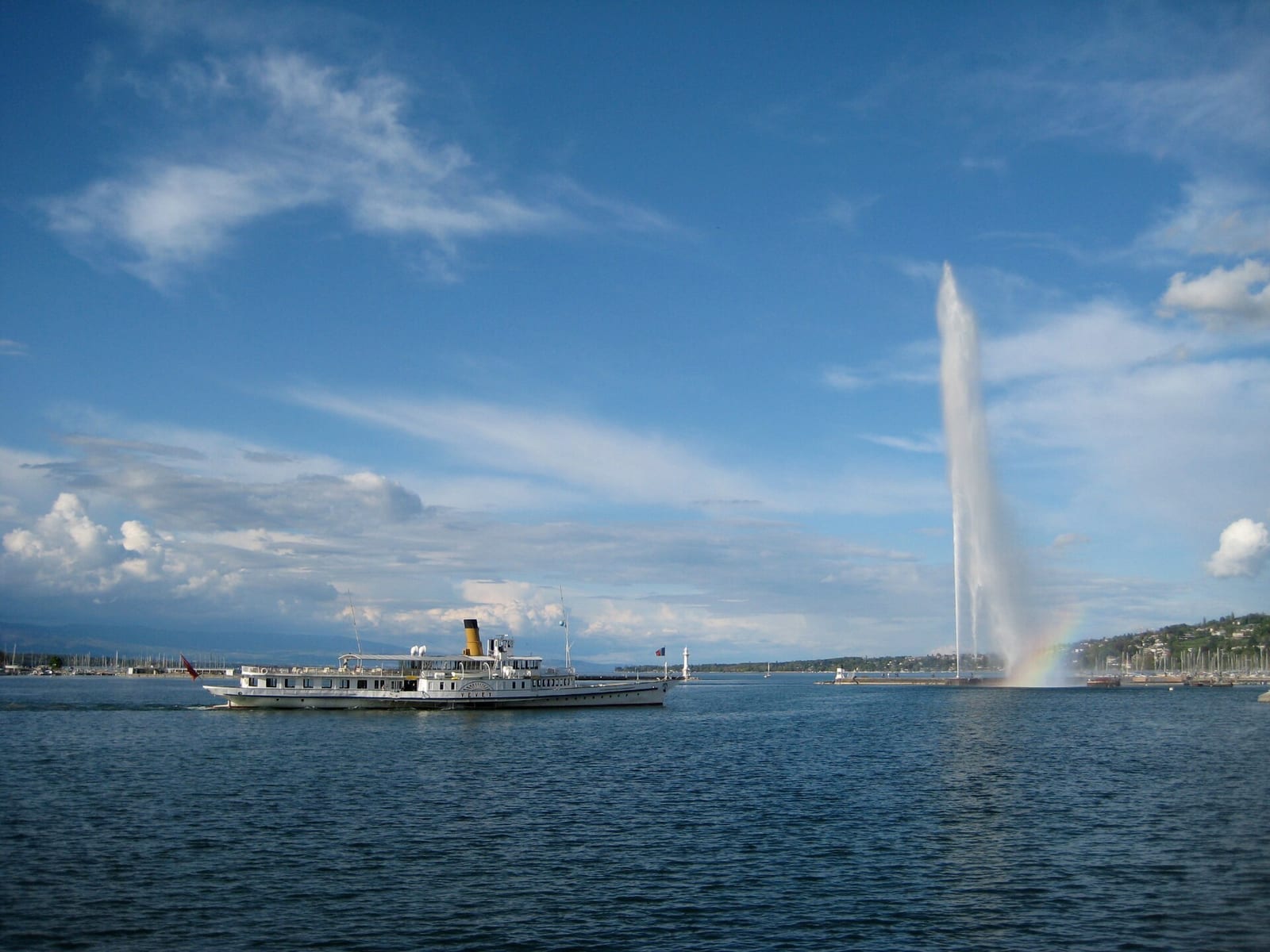 A paddle steamer passes the Jet d'Eau fountain on Lake Geneva under a blue sky, with the Geneva shoreline behind.