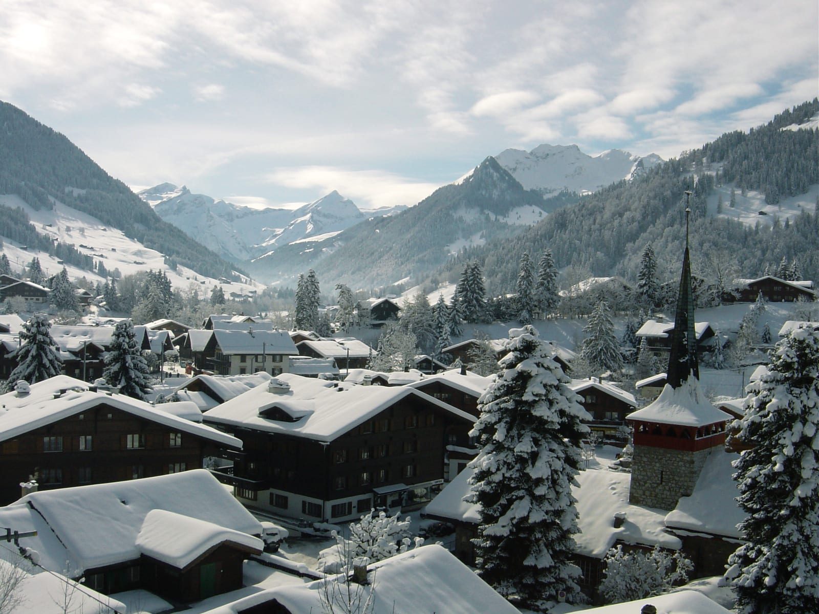 Gstaad village under fresh snow, chalet roofs and the church spire against forested slopes and the Saanenland peaks.