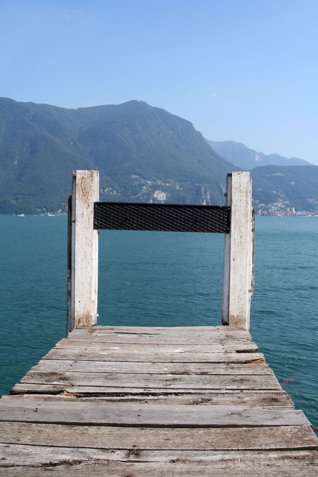 A wooden jetty leads out over the turquoise water of Lake Lugano toward the hills on the far shore.