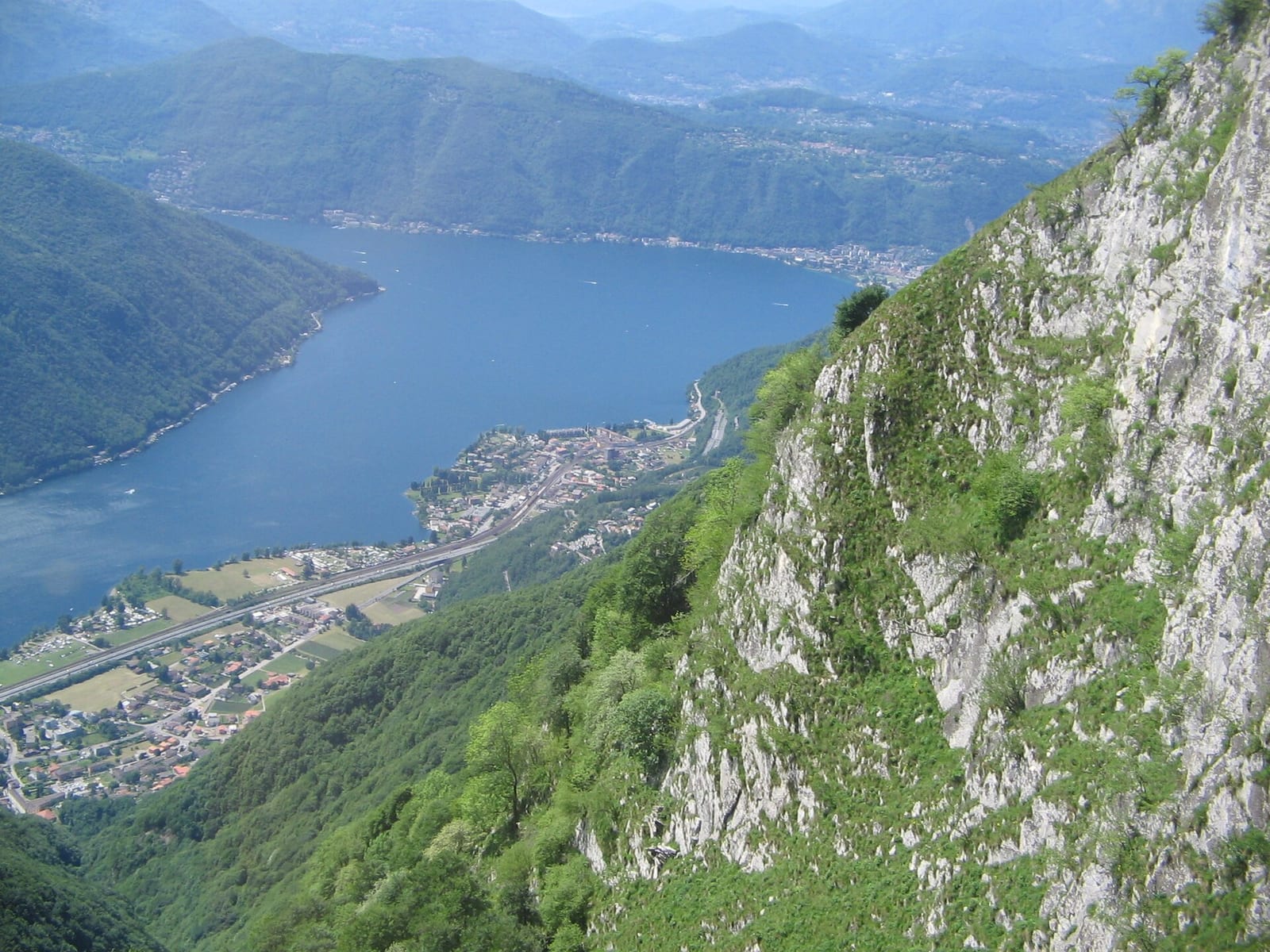 Lake Lugano and the town seen from the slopes of Monte Generoso, with green hillsides dropping to the water.