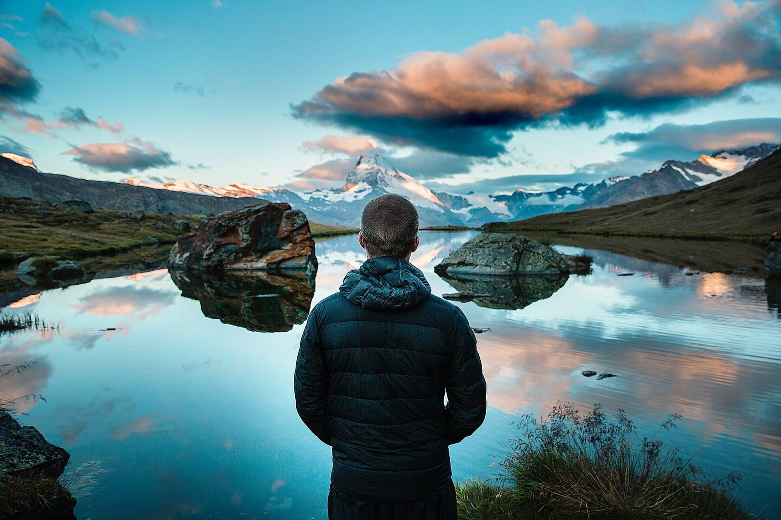 A person stands at a still mountain lake at sunrise as the Matterhorn and pink clouds reflect in the water.