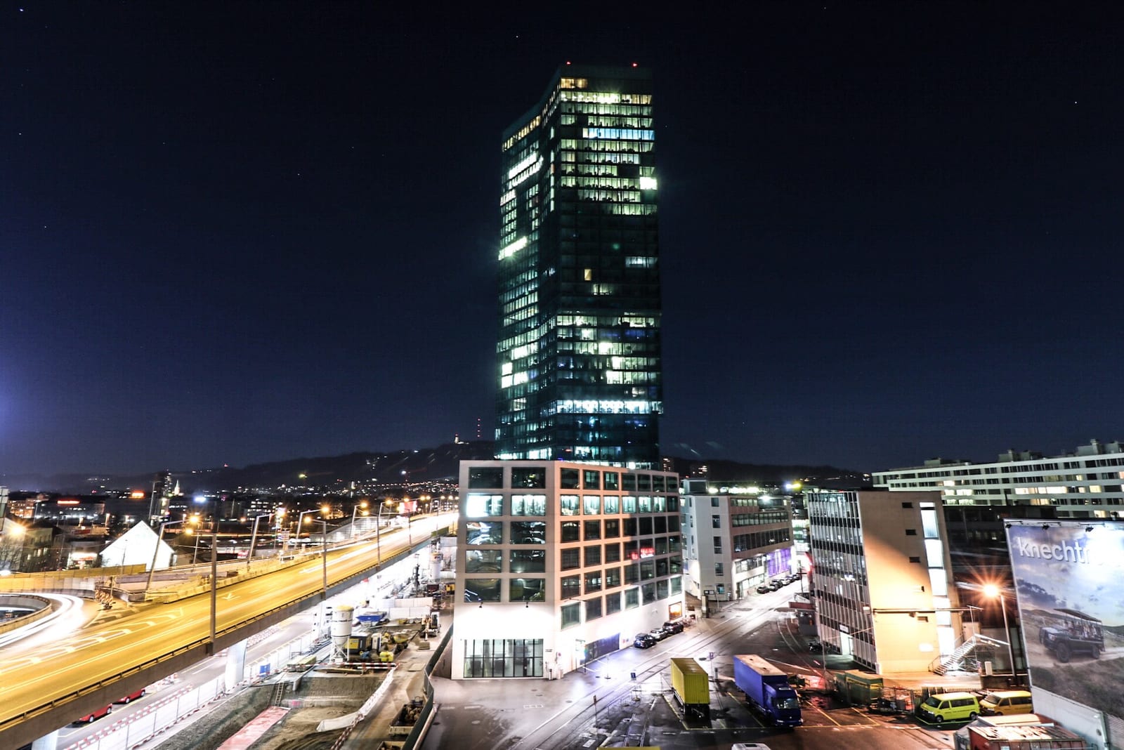 The dark-glass Prime Tower in Zürich-West lit at night above the Hardbrücke traffic and the surrounding office blocks.