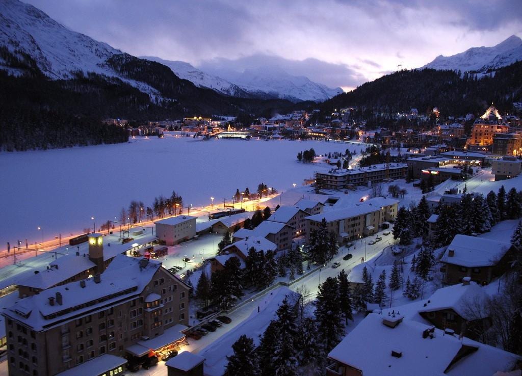 St. Moritz at dusk in winter, the lit town curving round the frozen lake with snow-covered Engadin mountains behind.