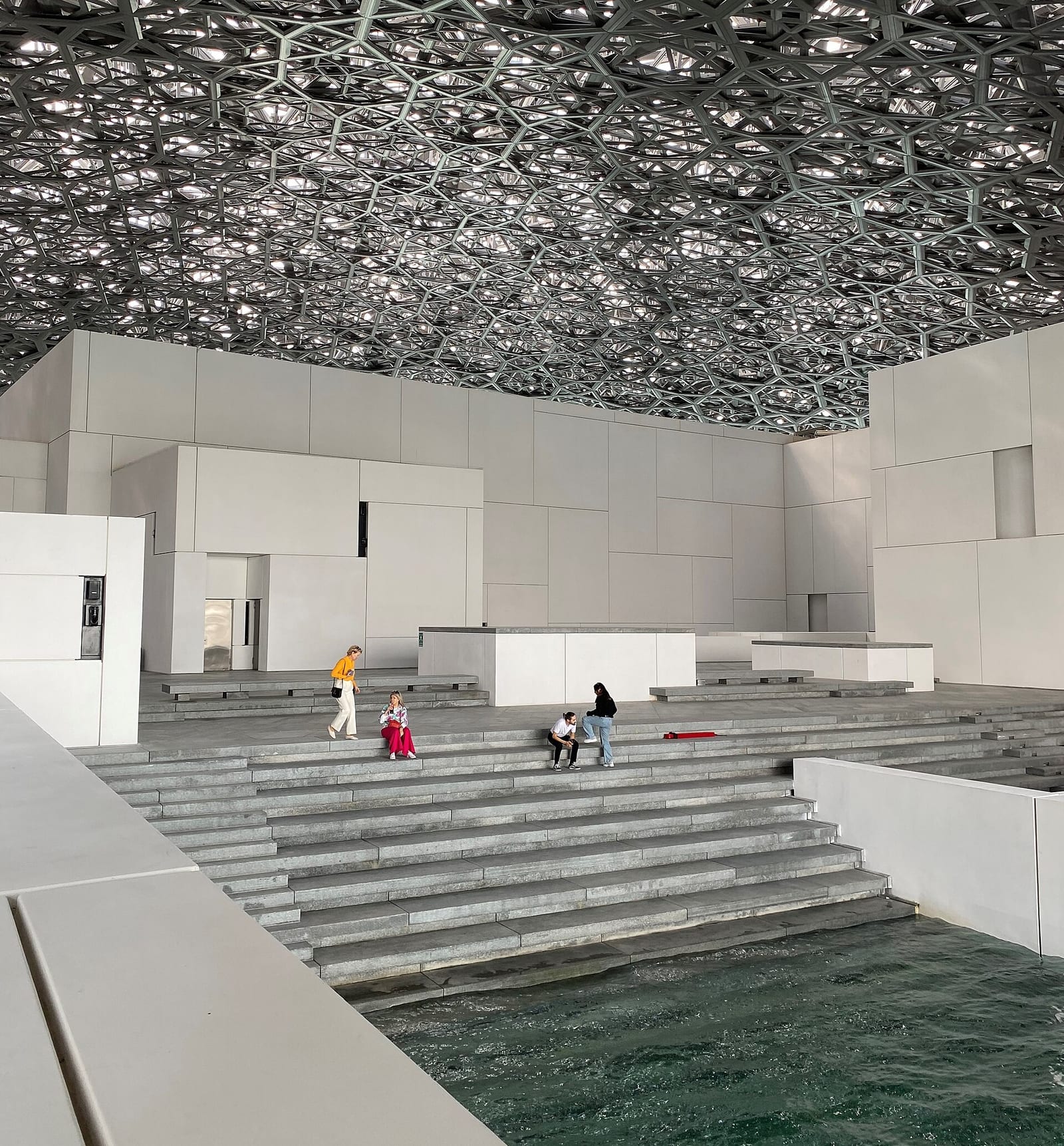 View under the perforated dome of the Louvre Abu Dhabi, dappled light falling on white walls and a pool of water.