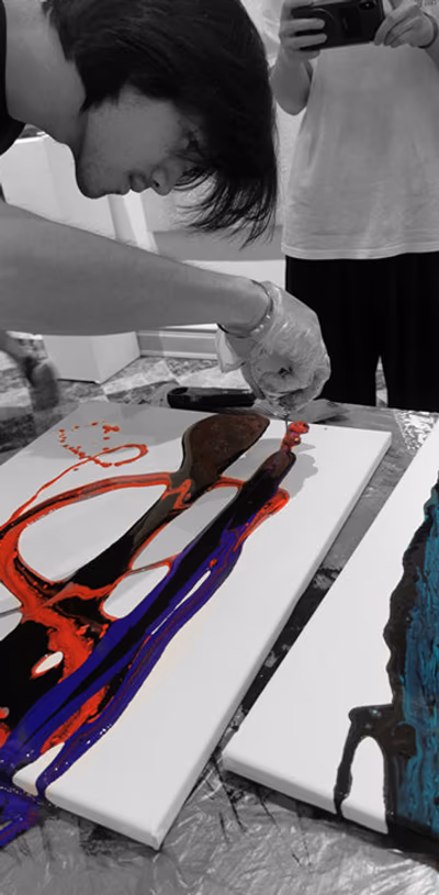 A painter pouring red and violet acrylic across a white canvas laid flat on the studio floor, photographed in black and white