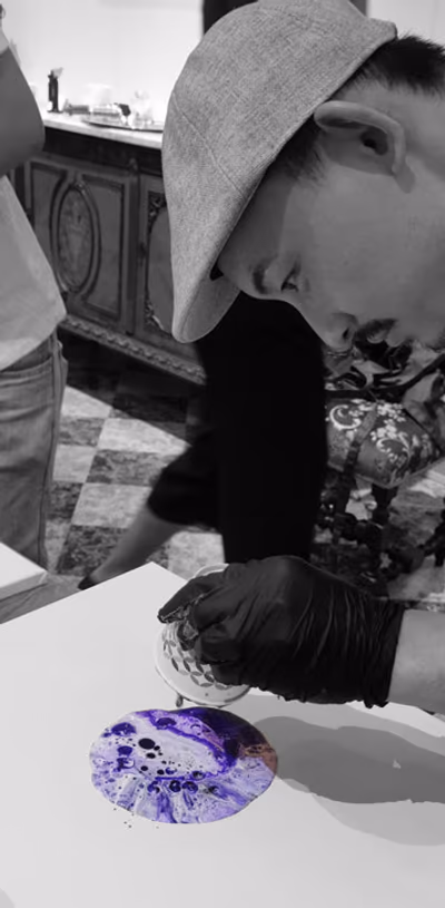 A painter in a felt hat tipping a cup of violet paint onto a fresh canvas, photographed in black and white